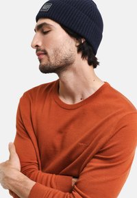 Pomarańczowy sweter z dzianiny o okrągłym dekolcie, z delikatnym akcentem logo. Granatowa czapka z prążkowanym wzorem dopełnia stylizację. Teksturowany materiał.