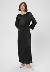 Frau in einem langen schwarzen Satin-Kleid mit gerafftem Detail an der Taille und seitlichem Schlitz, trägt beigefarbene spitze Pumps, steht vor einem schlichten Hintergrund.