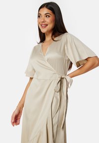 Robe portefeuille en satin beige avec de courtes manches flottantes, décolleté en V et un lien à la taille. Silhouette fluide avec une texture douce.