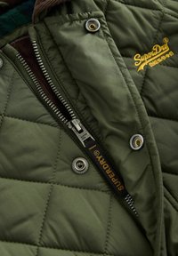 Olivza zaudējumus jaku ar quilted rakstu, metāla rāvējslēdzīgu, spiedpogu pogām un auduma etiķeti ar 'SuperDry' zīmolu tuvu rāvējslēdzēja.