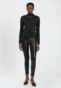 Mujer de pie contra un fondo blanco, vistiendo un cuello alto negro brillante, leggings de cuero negros y zapatos planos negros de punta.