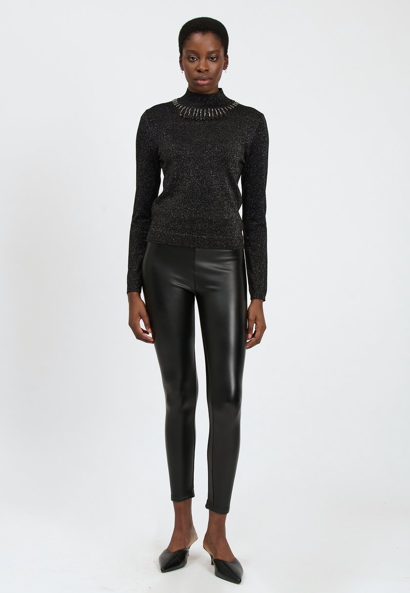 Donna in piedi su uno sfondo bianco, che indossa un dolcevita nero luccicante, leggings in pelle nera e ballerine a punta nere.