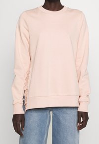 Roze sweatshirt van zachte stof met een ronde hals, lange mouwen en geplooide zijdetails. Gepaard met lichtblauwe jeans.