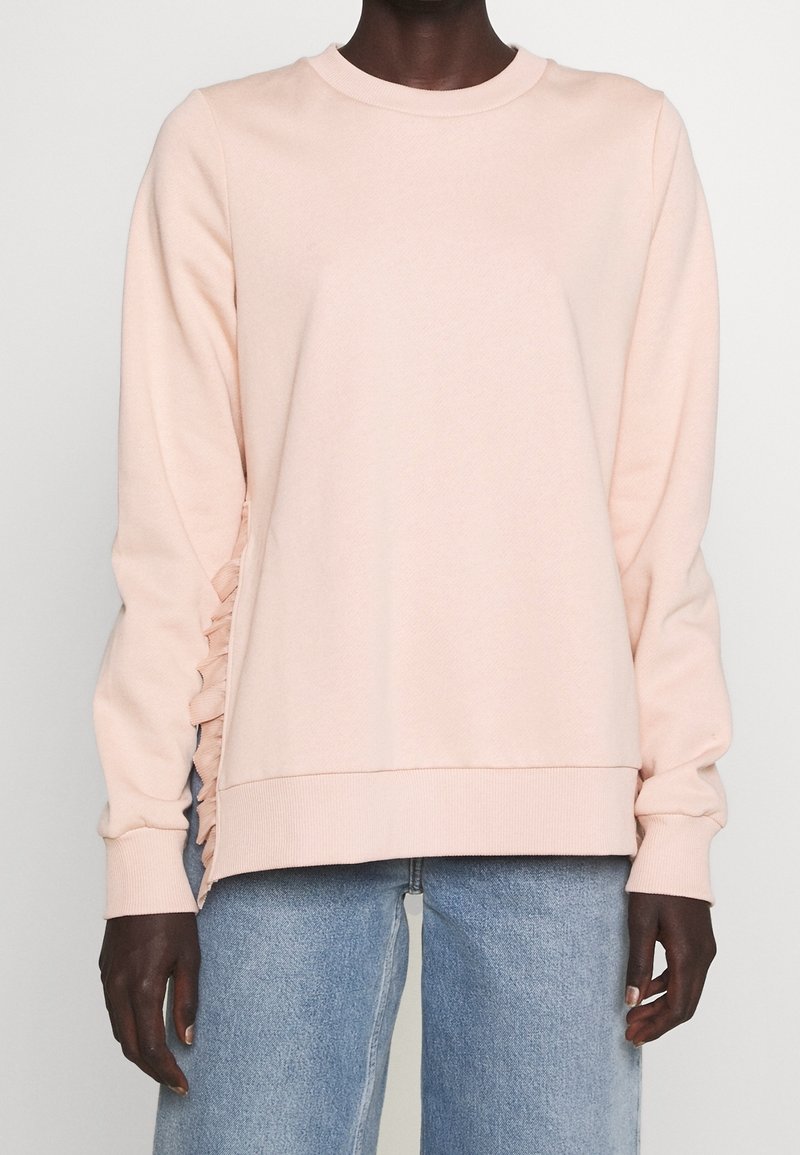 Roze sweatshirt van zachte stof met een ronde hals, lange mouwen en geplooide zijdetails. Gepaard met lichtblauwe jeans.