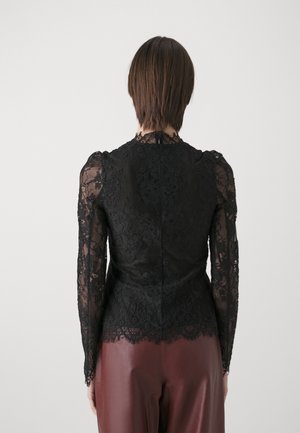 Woman with short brown hair wearing a black lace long-sleeve top and maroon leather pants, facing away against a plain background.