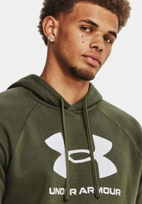 Felpa con cappuccio verde oliva con logo Under Armour bianco. Presenta stringhe e una texture morbida, progettata per comfort e durata.
