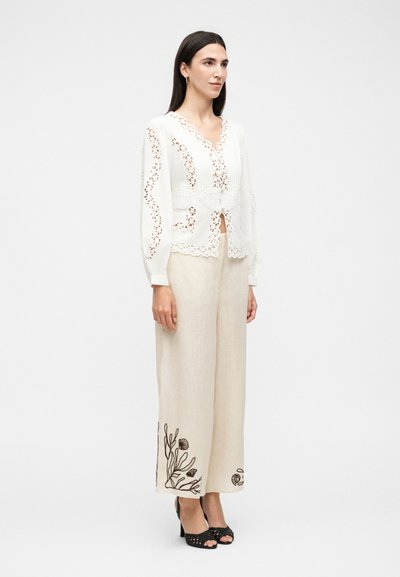 Γυναίκα που φοράει λευκό μπλουζάκι με μακριά μανίκια και δαντελωτές λεπτομέρειες, καθώς και μπεζ παντελόνι με φαρδιά πόδια και μαύρη κεντημένη λουλουδένια διακόσμηση στα τελειώματα, στέκεται με μαύρες γόβες.