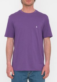 Camiseta corta de manga corta color púrpura, hecha de suave algodón, con un escote redondo y un pequeño logo blanco en el lado izquierdo del pecho. Se lleva con jeans azules.