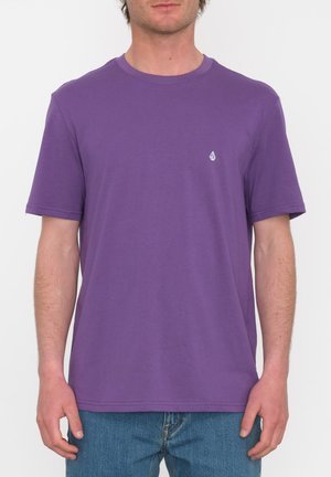 Camiseta corta de manga corta color púrpura, hecha de suave algodón, con un escote redondo y un pequeño logo blanco en el lado izquierdo del pecho. Se lleva con jeans azules.