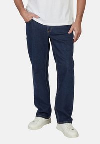 Hombre con jeans rectos de color azul oscuro y zapatillas blancas, con una mano en el bolsillo, contra un fondo liso.