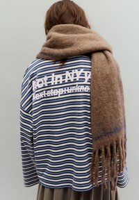 Cămașă în dungi albastru închis, alb și albastru deschis cu text imprimat; asortată cu un fular maro pufos, cu margini ciucurate.