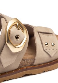 Sandal in suede beige con una grande fibbia ottagonale dorata, cuciture decorative e una soletta in sughero con piccoli chiodi in ottone lungo la suola.