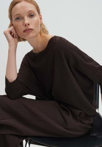 Blonde Frau trägt locker sitzendes dunkelbraunes Oberteil und Hose, sitzt auf einem schwarzen Stuhl, stützt ihren Kopf auf die Hand und sieht nachdenklich aus.