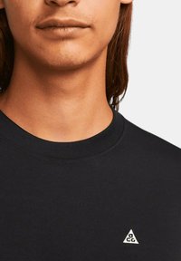 Svart bomulls t-shirt med rund halsringning, prydd med en liten vit triangellogo på bröstet. Slät textur och avslappnad passform.