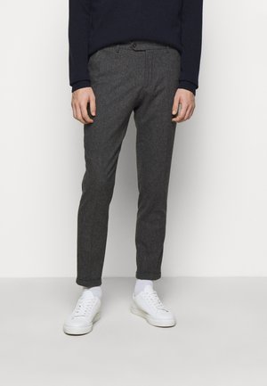 Homme portant un pantalon gris foncé slim fit, des chaussettes blanches, des baskets blanches et un pull bleu marine foncé, debout contre un mur clair uni.