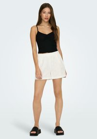 Débardeur noir à côtes avec des bretelles fines, associé à des shorts blancs amples avec des détails brodés et une taille élastique. Sandales noires à enfiler.