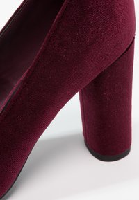 Chaussure à talon haut en velours bordeaux avec un talon carré épais et une surface en tissu lisse et texturée.