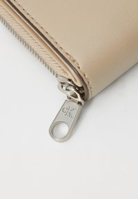 Portafoglio sintetico beige con una cerniera argentata caratterizzata dalle iniziali incise "CK" e una finitura testurizzata vicino alla zip.