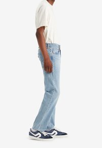 Homme portant un jean bleu clair, un t-shirt blanc et des baskets bleu marine et blanc, debout de profil sur un fond blanc.