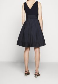 Nainen, joka on pukeutunut mustaan hihattomaan mekkoon, jossa on istuva vyötärö ja liehuva helma, yhdistettynä mustiin hihnakenkiin, joissa on läpinäkyvät korot.