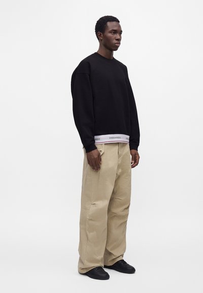 Homme debout portant un sweat-shirt noir, un pantalon beige à jambes larges et des chaussures noires sur un fond clair uni.