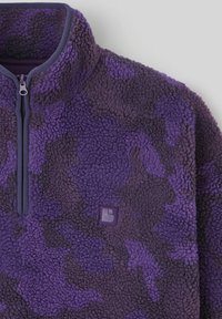 Sweat-shirt en polaire violet avec un col montant, fermeture éclair à moitié, et un motif de camouflage subtil. Présente un petit badge logo sur la poitrine.