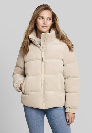 Junge Frau trägt eine beige Cord-Steppjacke mit Reißverschluss vorne und Händen in den Taschen, schaut seitlich vor einem einfarbigen Hintergrund.