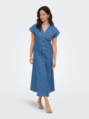 Mujer con un vestido largo azul con botones y mangas cortas, caminando con sandalias de tacón con tiras transparentes sobre un fondo liso.