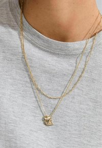 Gelaagde gouden kettingen met ingewikkeld textuurkettingen en een kleine driehoekige hanger met parels en accenten, gedragen over een grijs shirt.