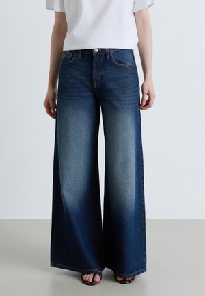 Femme portant un jean bleu taille haute à jambes larges, un haut blanc et des chaussures à bouts ouverts, debout devant un fond clair uni.