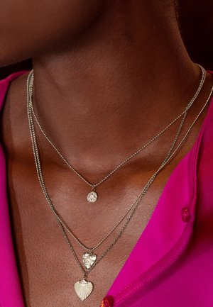 Drei silberne Halsketten, die übereinander liegend um den Hals getragen werden, mit einem strukturierten Herzanhänger, einem Blumenanhänger und einem gewellten Design. Über einem rosa Oberteil getragen.