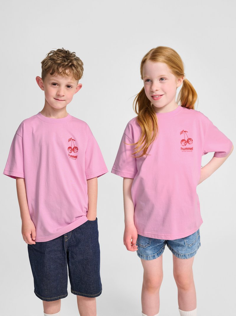 Dwoje dzieci stoi obok siebie, ubrane w pasujące różowe koszulki z czerwonym logo wiśni, połączone z dżinsowymi szortami i swobodnymi pozami.
