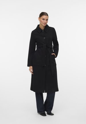 Frau trägt einen schwarzen, taillierten langen Mantel über dunkler Hose und schwarzen spitzen Schuhen, steht mit einer Hand in der Tasche vor einem weißen Hintergrund.