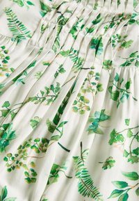 Kukallinen mekko, jossa on vihreitä lehtikuvioita valkoisella taustalla. Pehmeä kangas kerättyine tekstuureineen korostaa luonnosta inspiroitua muotokieltä.