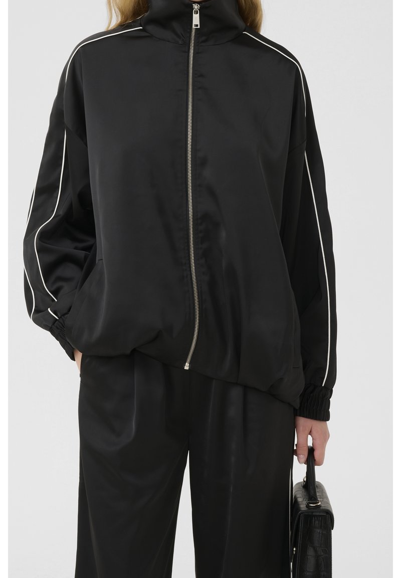 Personne portant une veste noire zippée avec un liseré blanc sur les manches et un pantalon noir assorti, tenant un sac à main noir texturé.