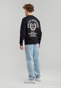Black sweatshirt with "ATHLETIC DEPARTMENT" text, crew neck, and graphic print on back; paired with light blue jeans and white sneakers.