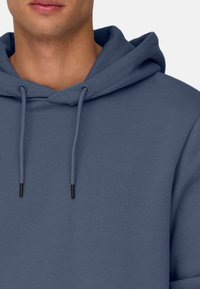 Felpa con cappuccio di un blu medio, realizzata in un tessuto morbido con una superficie liscia. Presenta cordini con punte nere e una vestibilità comoda.