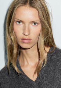 Blonde woman with light green eyes wearing a dark gray V-neck sweater, looking directly at the camera against a white background.