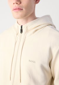 Nærbillede af en person iført en cremet lynlås-hættetrøje med en sort lynlås og "BOSS" logo på brystet.