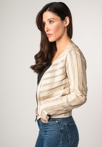 Giacca bomb bomber cropped in pelle beige con texture a righe orizzontali, chiusura con zip e dettagli cuciti, abbinata a jeans blu.