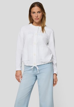 Woman wearing a white long-sleeve buttoned jacket with front pockets and light blue pants against a plain light gray background.