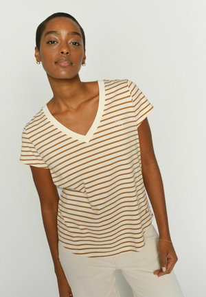 Frau mit kurzen Haaren, die ein beige-braun gestreiftes V-Ausschnitt-T-Shirt und hellfarbene Hosen trägt, vor einem einfarbigen Hintergrund.