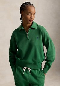 Femme aux cheveux tressés portant un sweat à capuche vert zippé et un pantalon vert assorti avec cordon de serrage, regardant vers le bas les mains dans les poches.