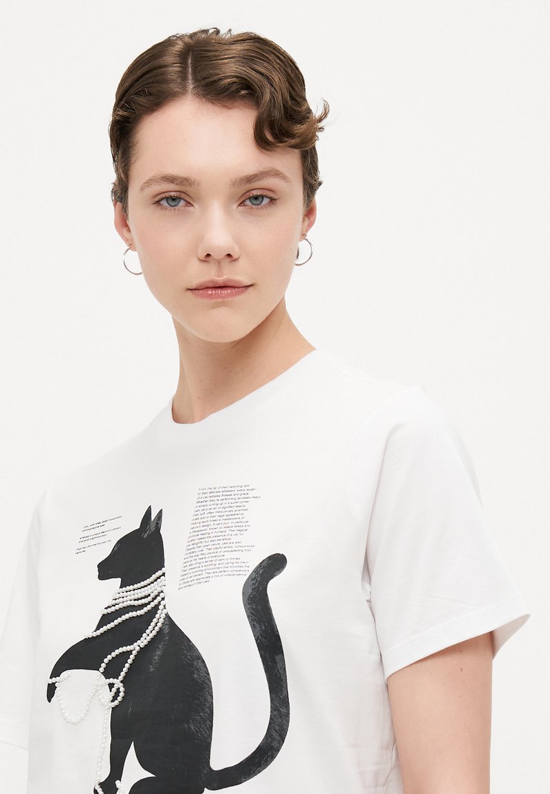 Weißes T-Shirt mit einem schwarzen Katzenmotiv und einem Design einer Perlenkette. Kurze Ärmel und runder Halsausschnitt. Text in einer verspielten Schriftart an der Seite.