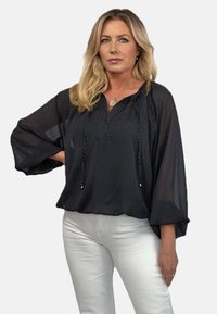 Czarna bluzka w białe kropki, luźny krój, okrągły dekolt i rękawy o długości trzy czwarte. Noszona z białymi spodniami.