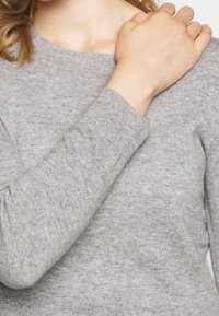 Personne portant un pull en maille gris clair, la main droite reposant sur l'épaule gauche, sur un fond neutre.