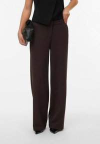 Vida byxor i mörkbrunt tyg med en slät textur, i kombination med en svart topp och en liten svart clutch. Spetsiga klackskor syns.