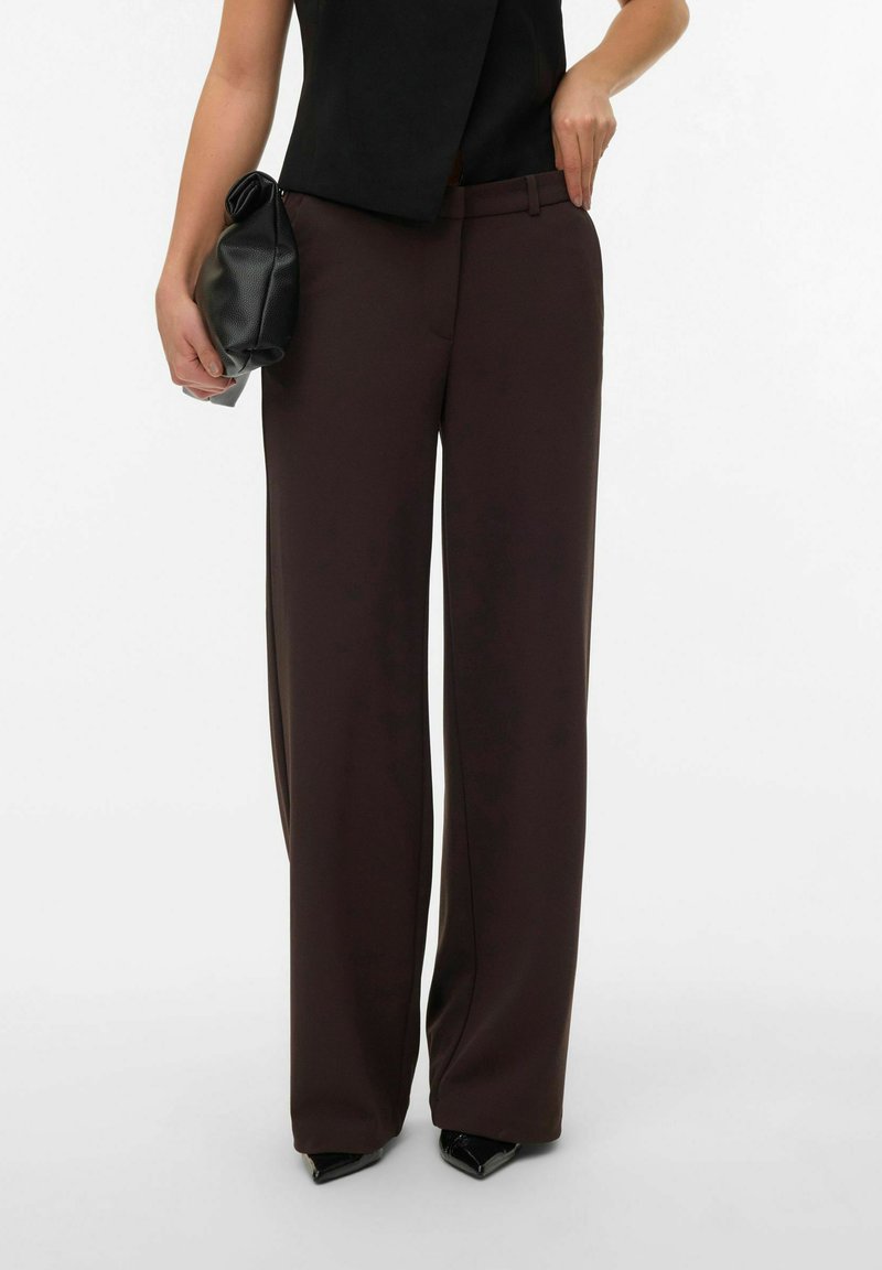 Vida byxor i mörkbrunt tyg med en slät textur, i kombination med en svart topp och en liten svart clutch. Spetsiga klackskor syns.
