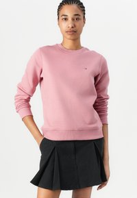 Pink sweatshirt made of soft fabric, featuring a crew neckline and ribbed cuffs. Notable small logo in contrasting colors on the chest. Black pleated skirt.