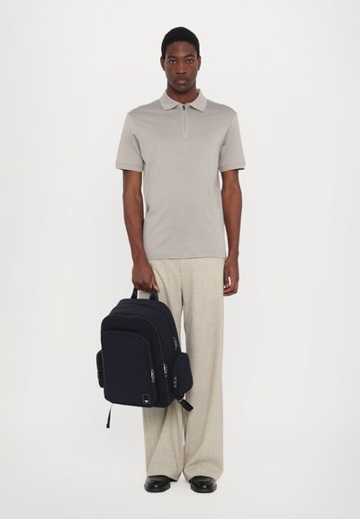 Γκρι πόλο μπλούζα με γιακά, συνδυασμένη με ανοιχτόχρωμα παντελόνια καρό. Το μοντέλο κρατάει μια σκούρα μπλε τσάντα πλάτης με πολλές θήκες και φερμουάρ.